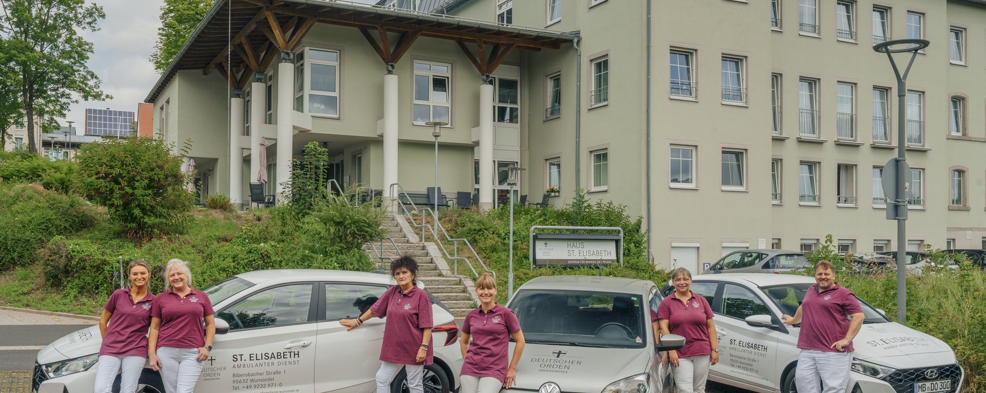 Vor einem Gebäudeeingang stehen zwei Fahrzeuge des Pflegedienstes Wunsiedel. Im Vordergrund gehen sechs Mitarbeiterinnen in orangenen T-Shirts lächelnd auf den Betrachter zu.